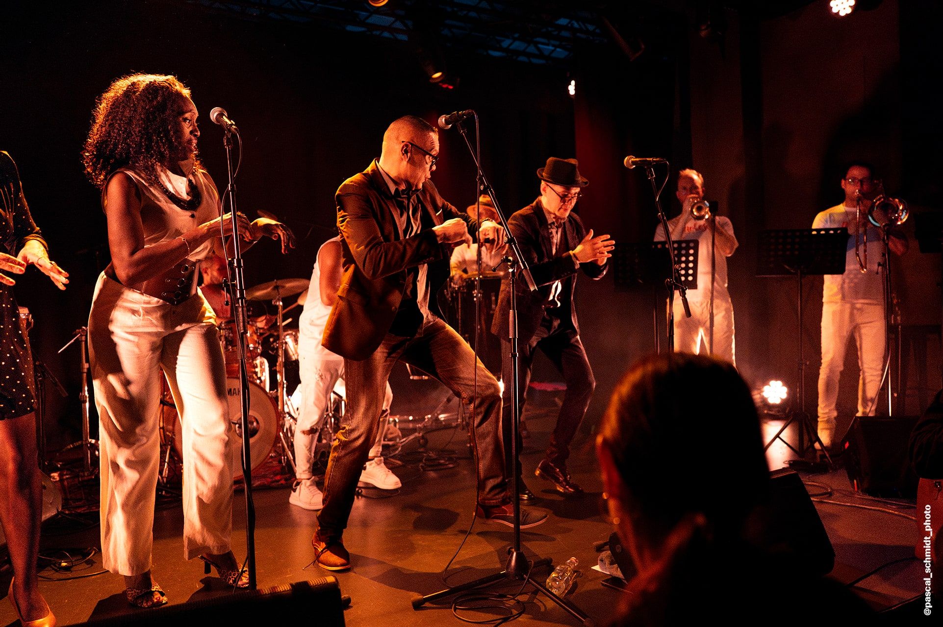 Les chanteurs de Nolosé dansent en front de scène dans la lumière ambrée d'un club, cuivres et batterie derrière eux — photo Pascal Schmidt