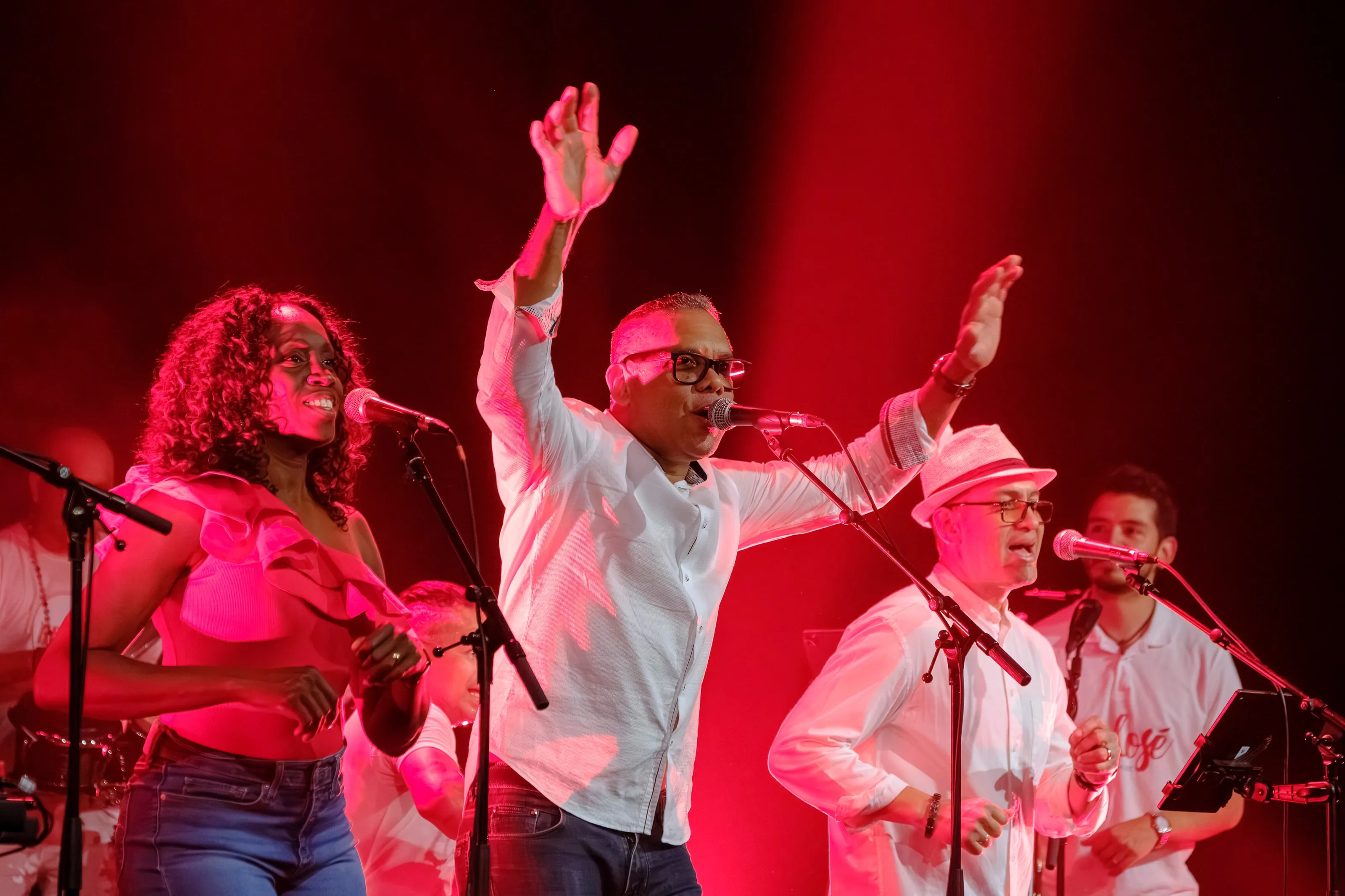 Les chanteurs lèvent les bras dans une lumière rouge intense, l'orchestre en blanc derrière eux
