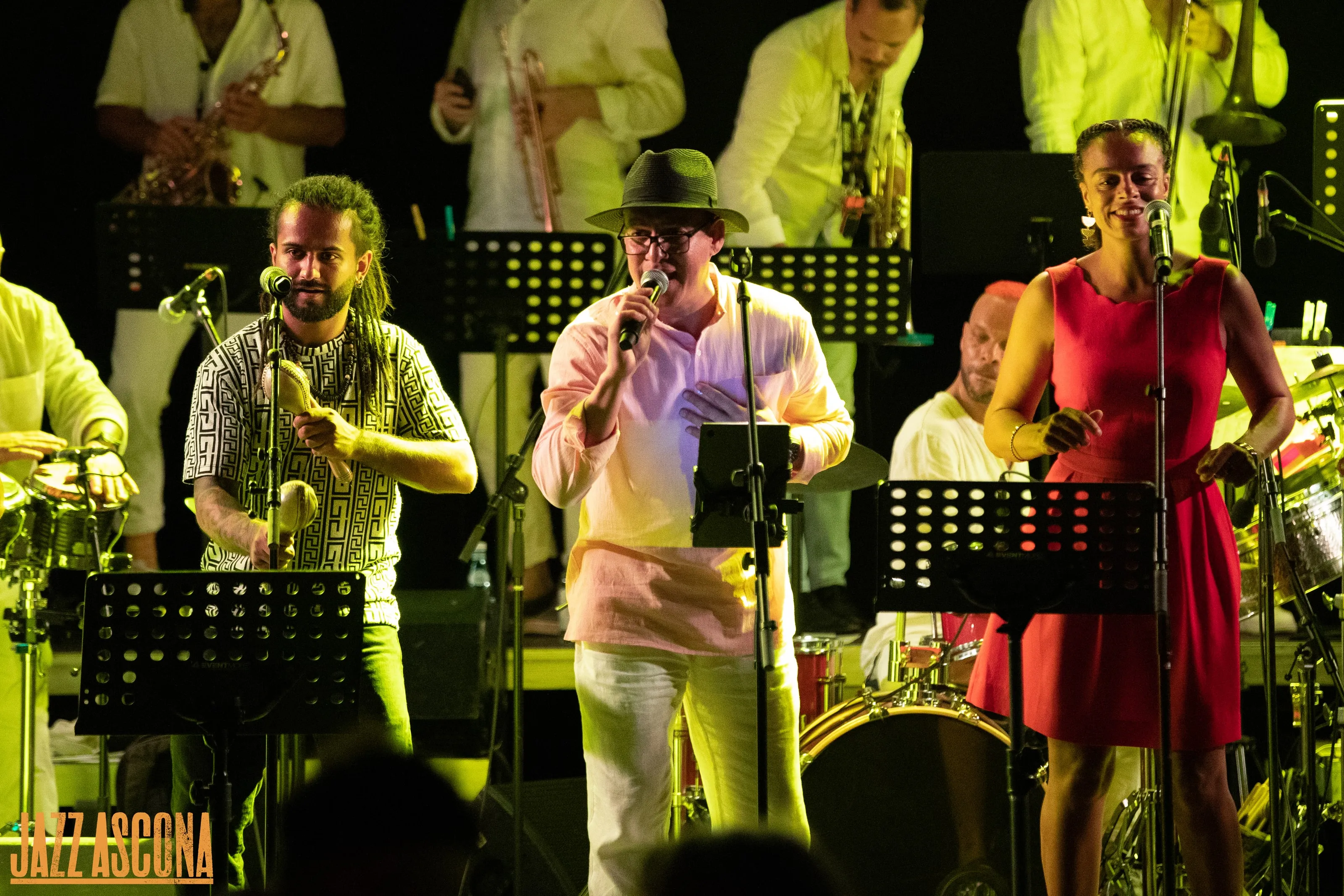 Trois chanteurs en front de scène dans une lumière ambrée à JazzAscona, les cuivres alignés derrière eux